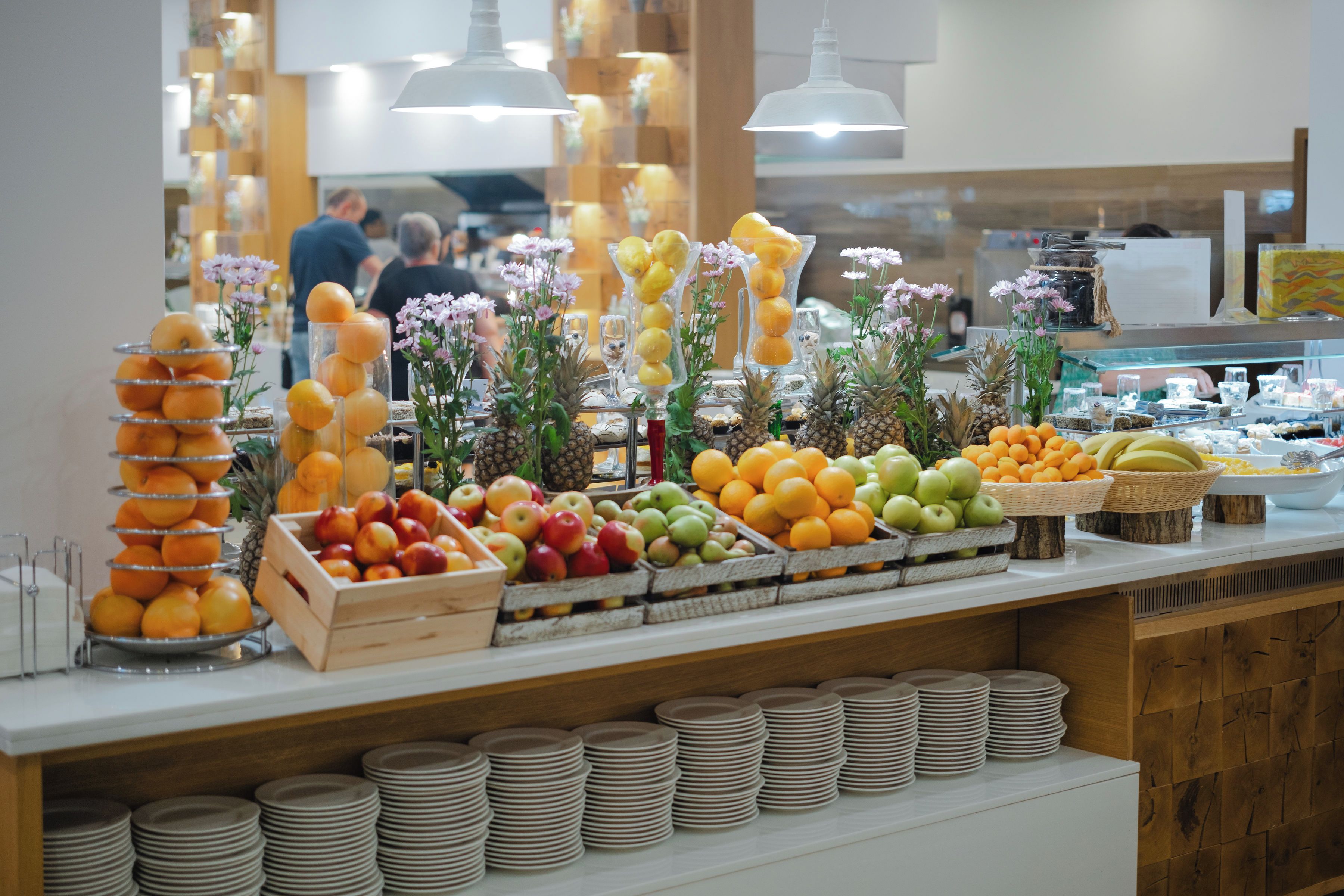 A buffet with various fruits and vegetables, including oranges, apples, and bananas, arranged on a counter with plates and bowls.