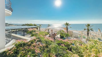 A balcony with a view of the sea, a beach with umbrellas, and a building with solar panels.