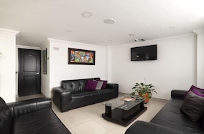 Una sala de estar moderna con un sofá de cuero negro, una mesa de centro de vidrio, un televisor de pantalla plana montado en la pared y una puerta que da a otra habitación.
