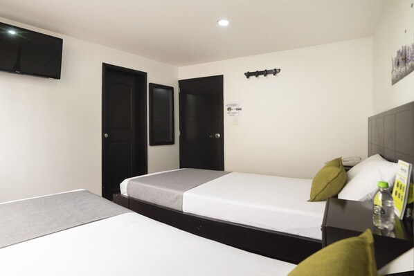 A hotel room with two beds, a flat-screen TV, a bedside table with a bottle, and a wall-mounted light.
