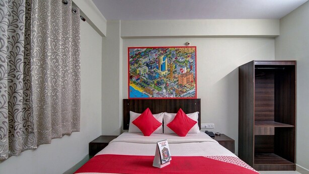 Habitación de hotel con una cama grande, dos almohadas rojas, una manta roja, una mesita de noche con un teléfono, y un cuadro de un paisaje urbano colorido sobre la cama.