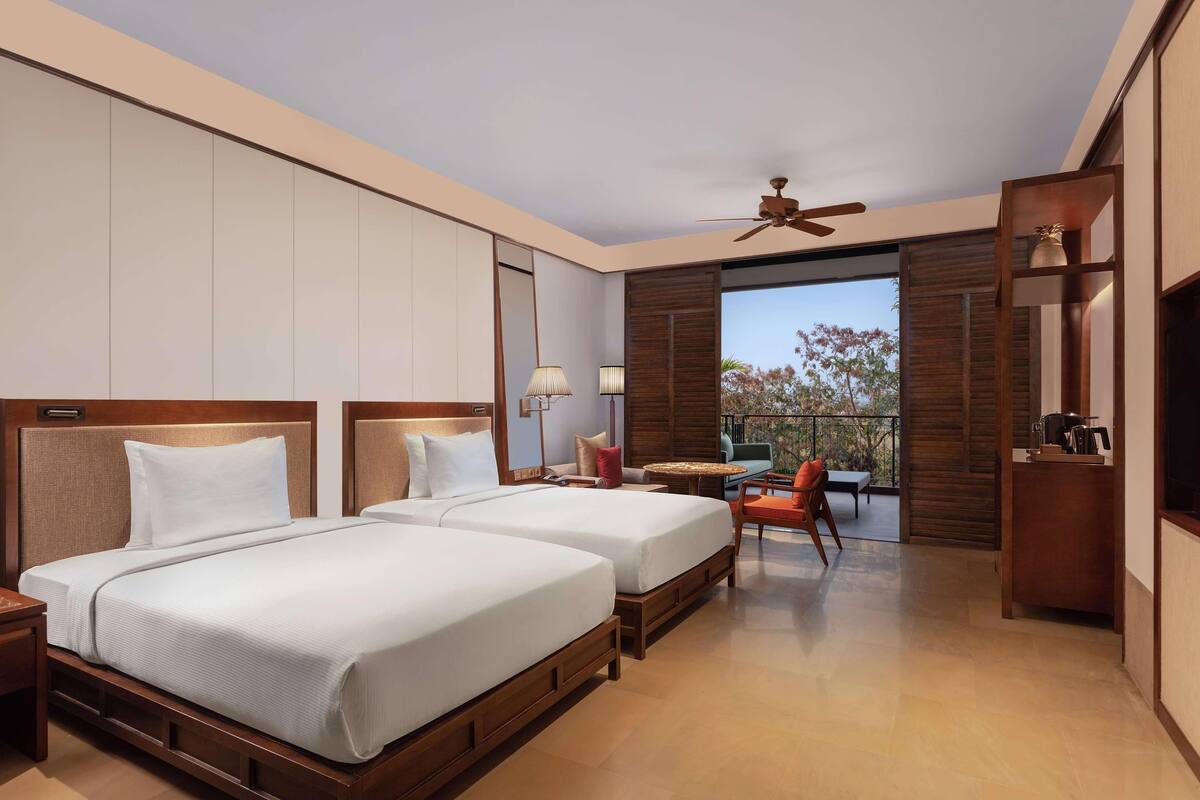 A hotel room with two beds, a balcony, a ceiling fan, a small table, and a TV.