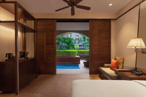A hotel room with a bed, a desk with a coffee maker, a lamp, a ceiling fan, and a view of a pool and outdoor seating area.