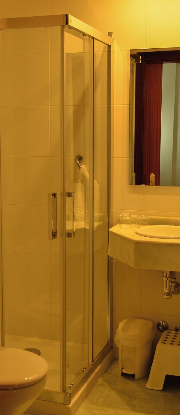 A bathroom with a glass shower enclosure, a toilet, a sink with a mirror above it, and a small stool.
