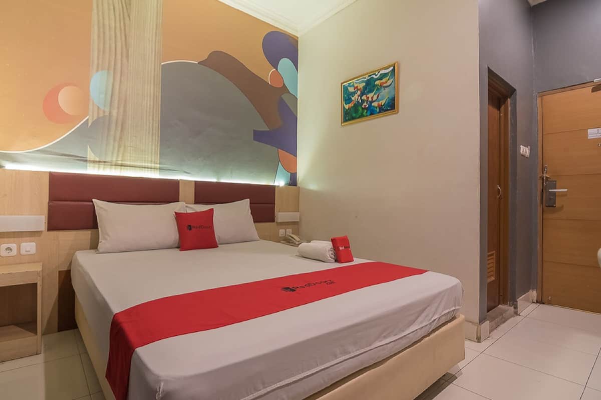 A hotel room with a bed, a nightstand, a red throw, a painting, and a wooden door.