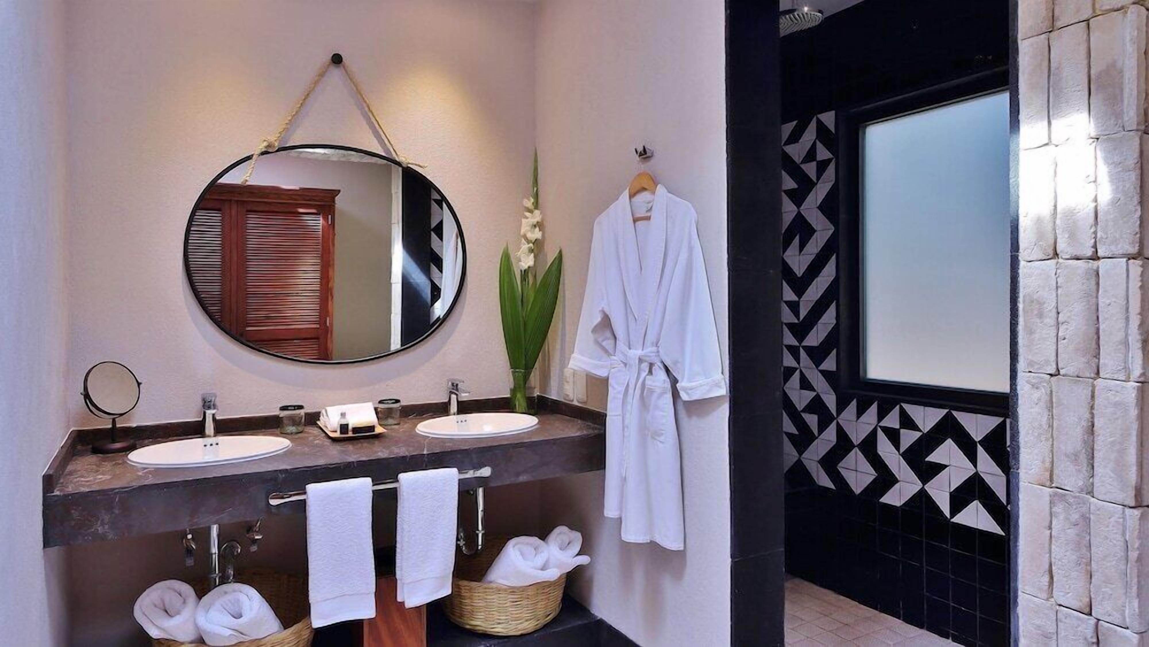 A bathroom with two sinks, a round mirror, and a robe hanging on the wall.