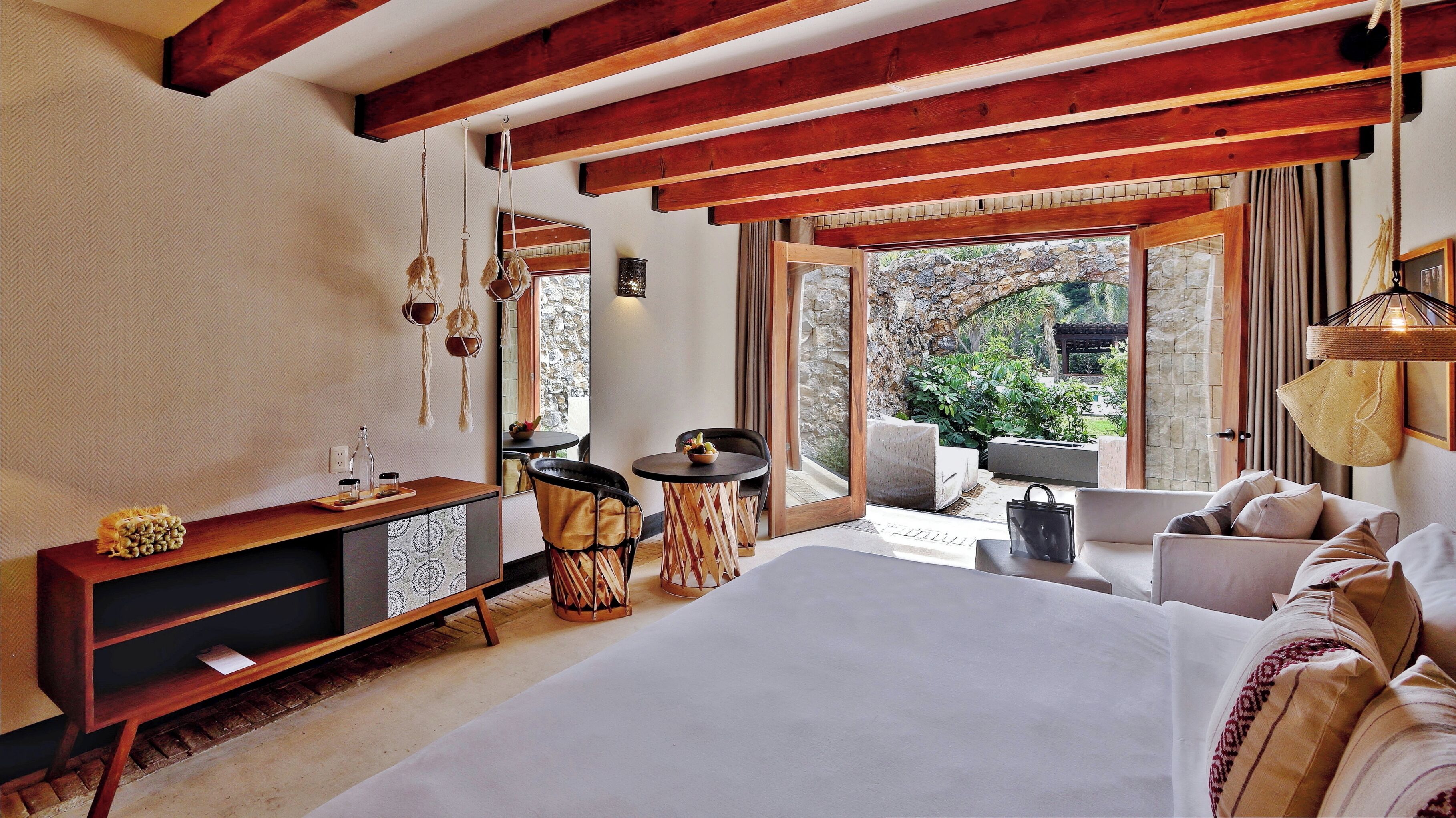 A bedroom with a large bed, a wooden desk, a round table, and a sofa. There is a view of greenery outside through large windows.