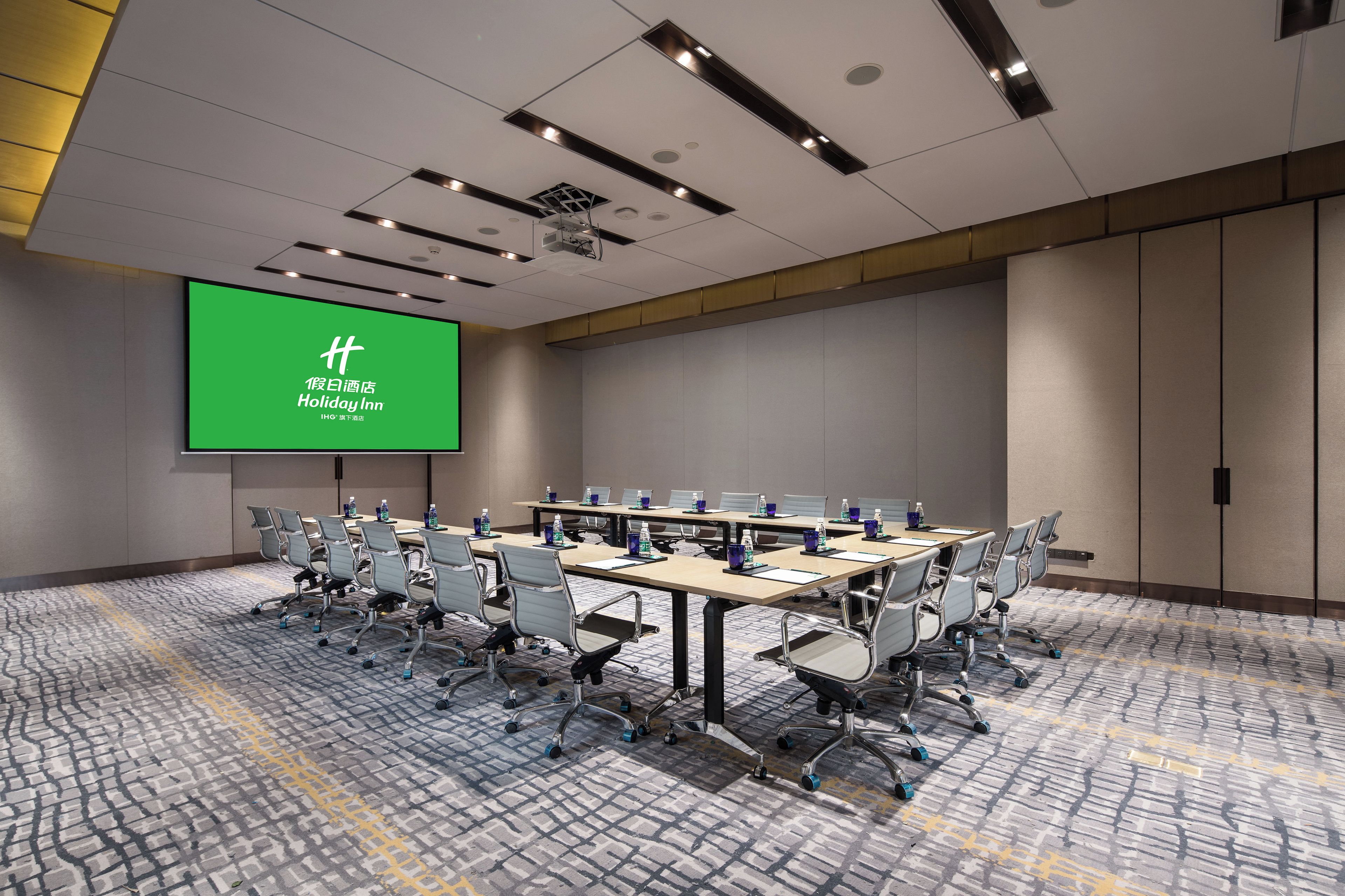 Ein Konferenzraum mit einer großen Leinwand, auf der ein Hotel-Logo zu sehen ist, mehreren Tischen mit Stühlen und einem gemusterten Teppich.