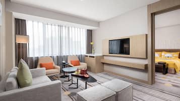 A modern hotel room with a sofa, armchair, coffee table, TV, and a bed with a yellow bedspread.