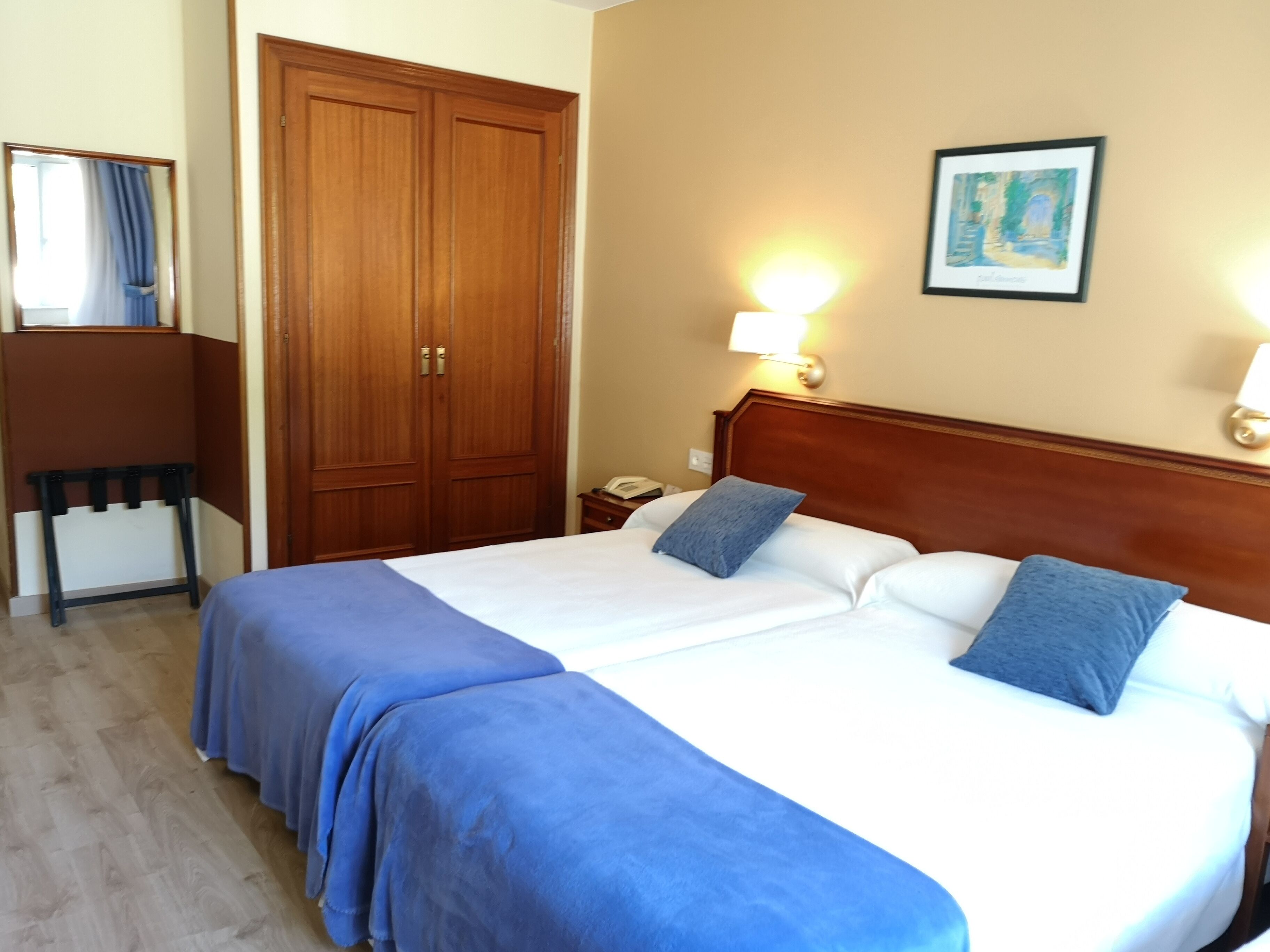 Una cama doble con ropa de cama azul, un armario de madera, una mesita de noche con una lámpara y un cuadro enmarcado en la pared.