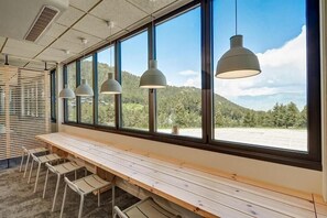 Eine lange Holzbank mit Metallstühlen, große Fenster mit Blick auf einen bewaldeten Hang und Pendelleuchten.