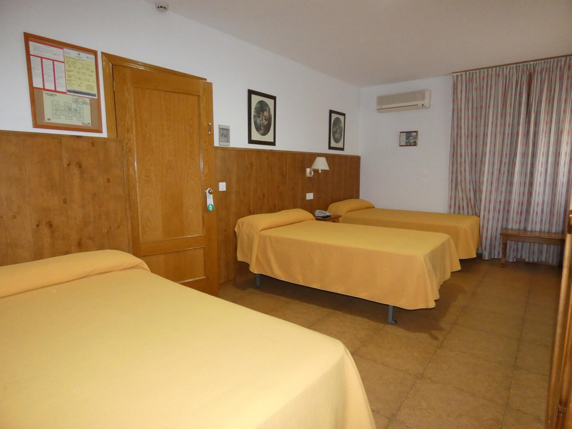 Habitación de hotel con tres camas individuales, un armario de madera, aire acondicionado instalado en la pared y una cortina.