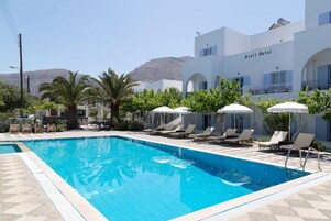 Una piscina con sillas de descanso y sombrillas, un edificio de hotel con el letrero "Aroti Hotel" y una montaña al fondo.