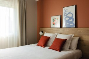A hotel room with a bed, two pillows, a headboard, a wall with artwork, and a window with curtains.
