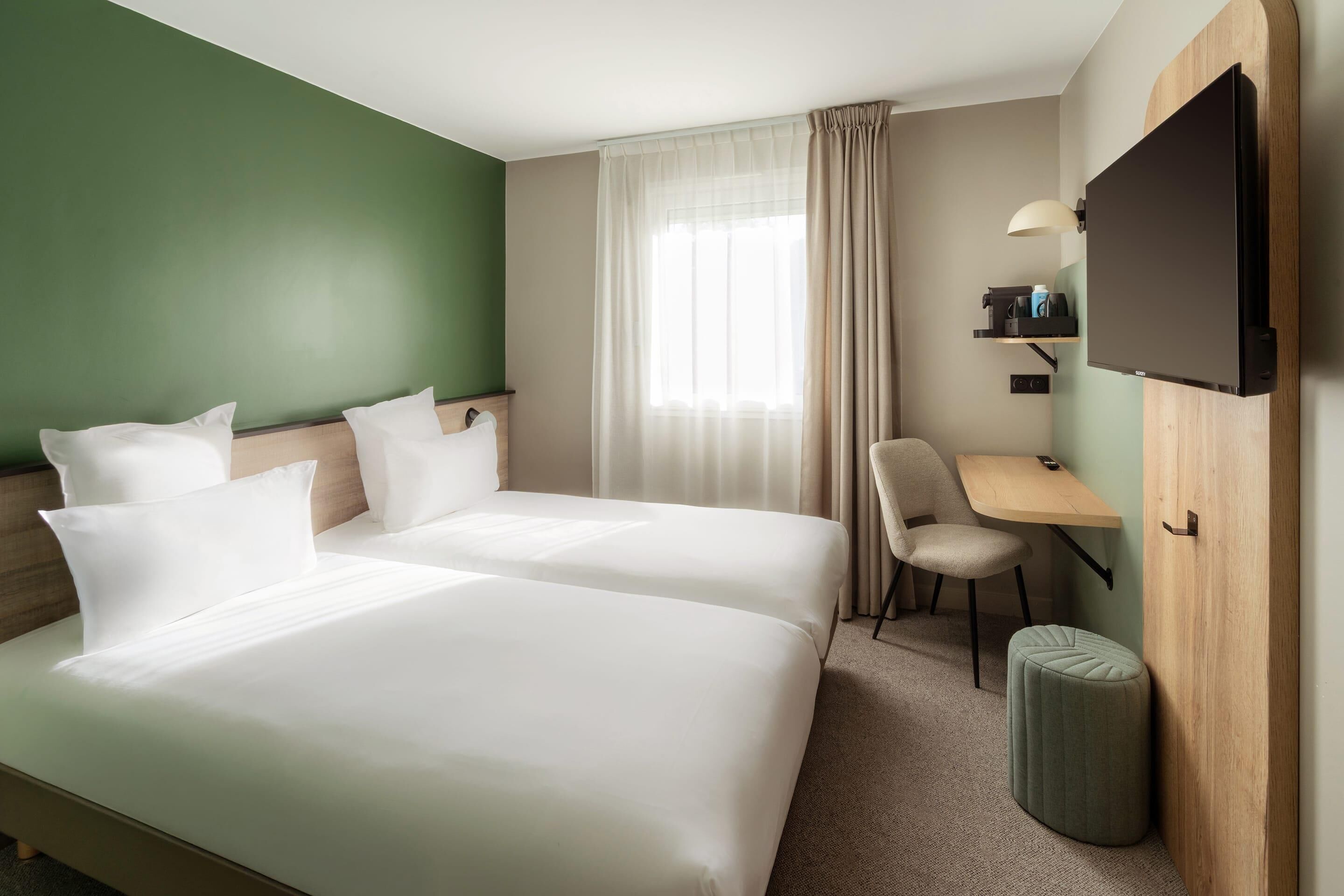 A hotel room with two beds, a desk, a chair, a TV, and a small ottoman.
