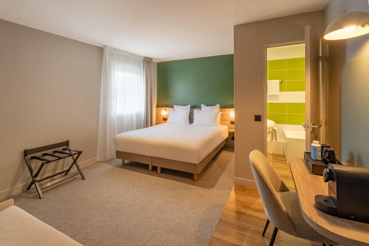 A hotel room with a bed, a desk with a coffee machine, a chair, a small table, and a bathroom visible through an open door.