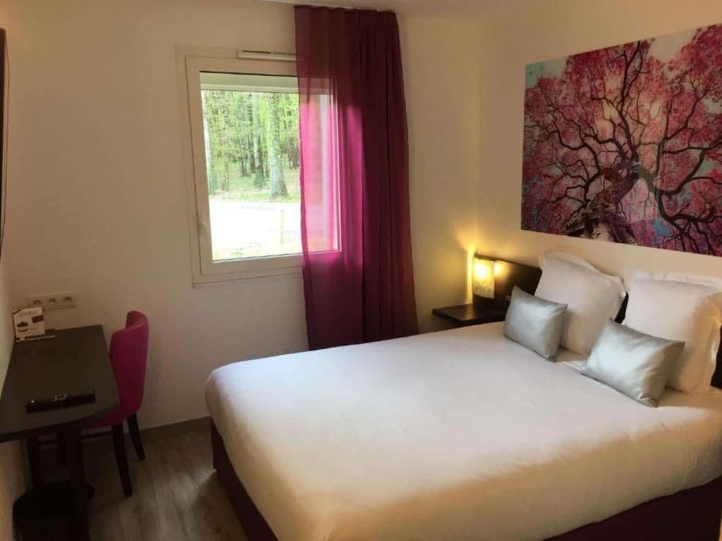 Una habitació d'hotel amb un llit, escriptori, cadira, finestra amb cortines i un quadre al mur.