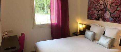 A hotel room with a bed, a desk, a chair, a window with curtains, and a painting on the wall.