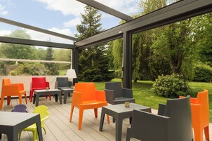 A covered outdoor seating area with tables and chairs, a lamp, and a view of a garden.