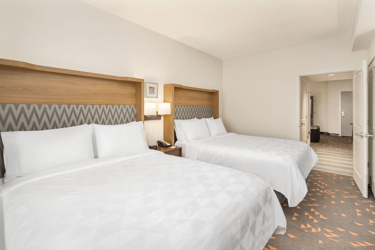 Habitación de hotel con dos camas, una mesita de noche con lámpara y una puerta que da a otra habitación.
