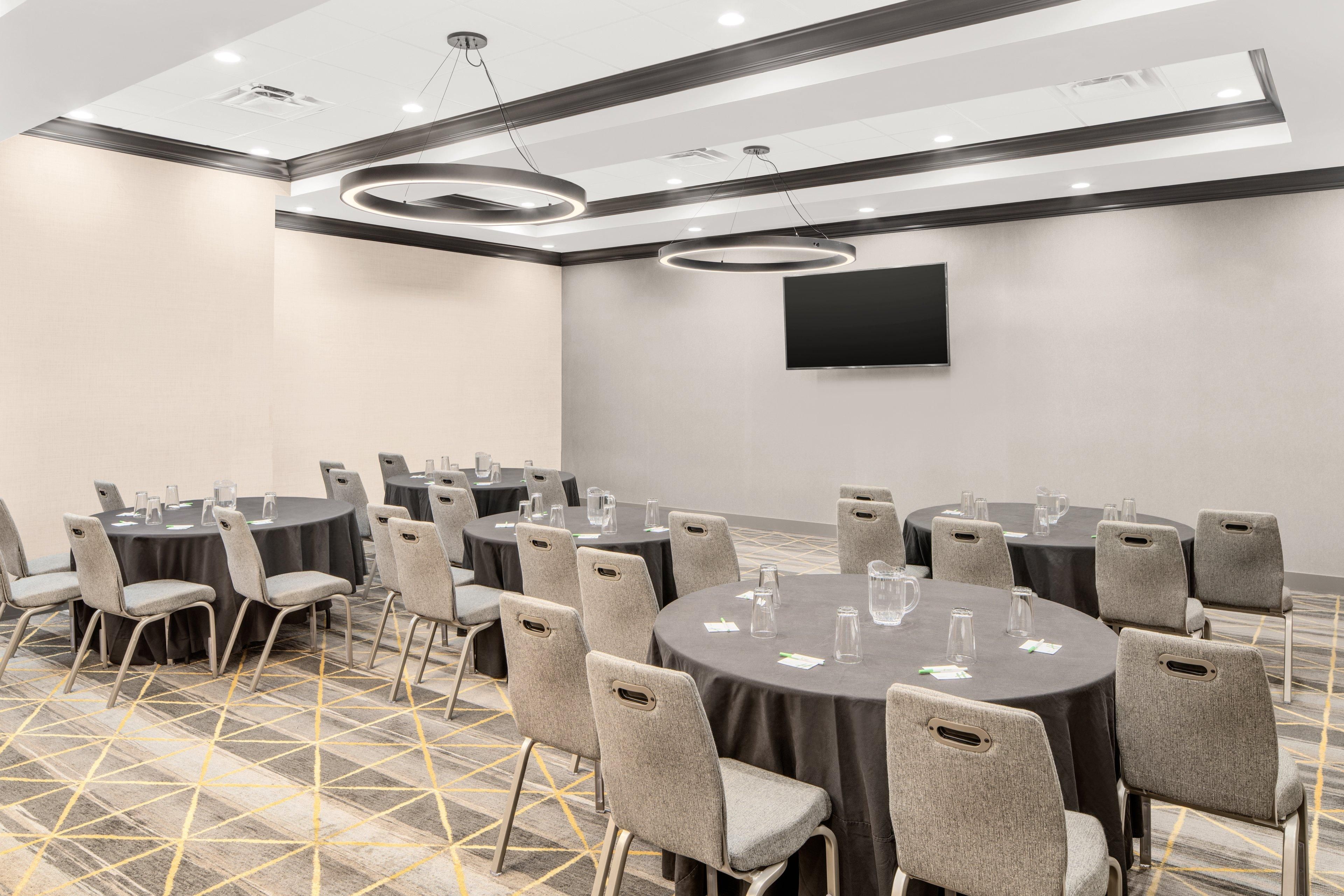 Une salle de conférence avec des tables rondes, des chaises grises, un téléviseur à écran plat et une moquette à motifs.