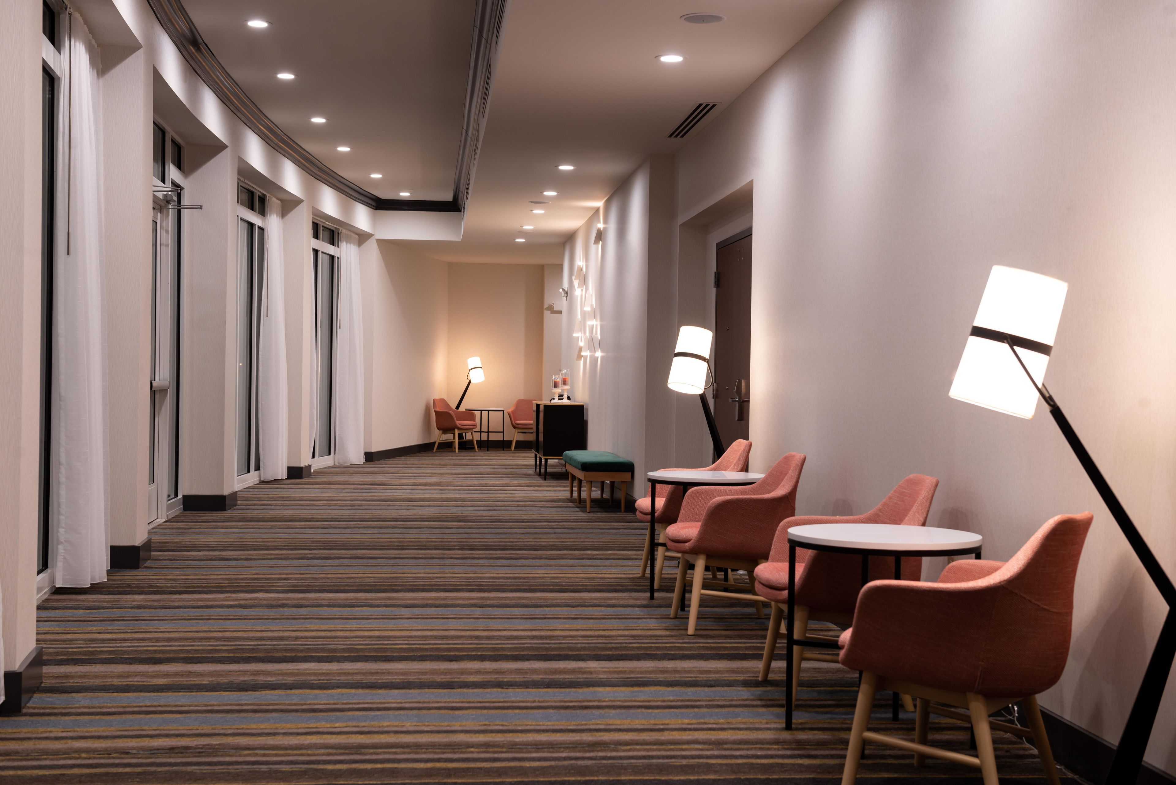Un couloir d’hôtel avec un sol recouvert de moquette, un espace détente avec des chaises et des tables, et un comptoir d’accueil.
