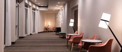 A hotel corridor with a carpeted floor, seating area with chairs and tables, and a reception desk.