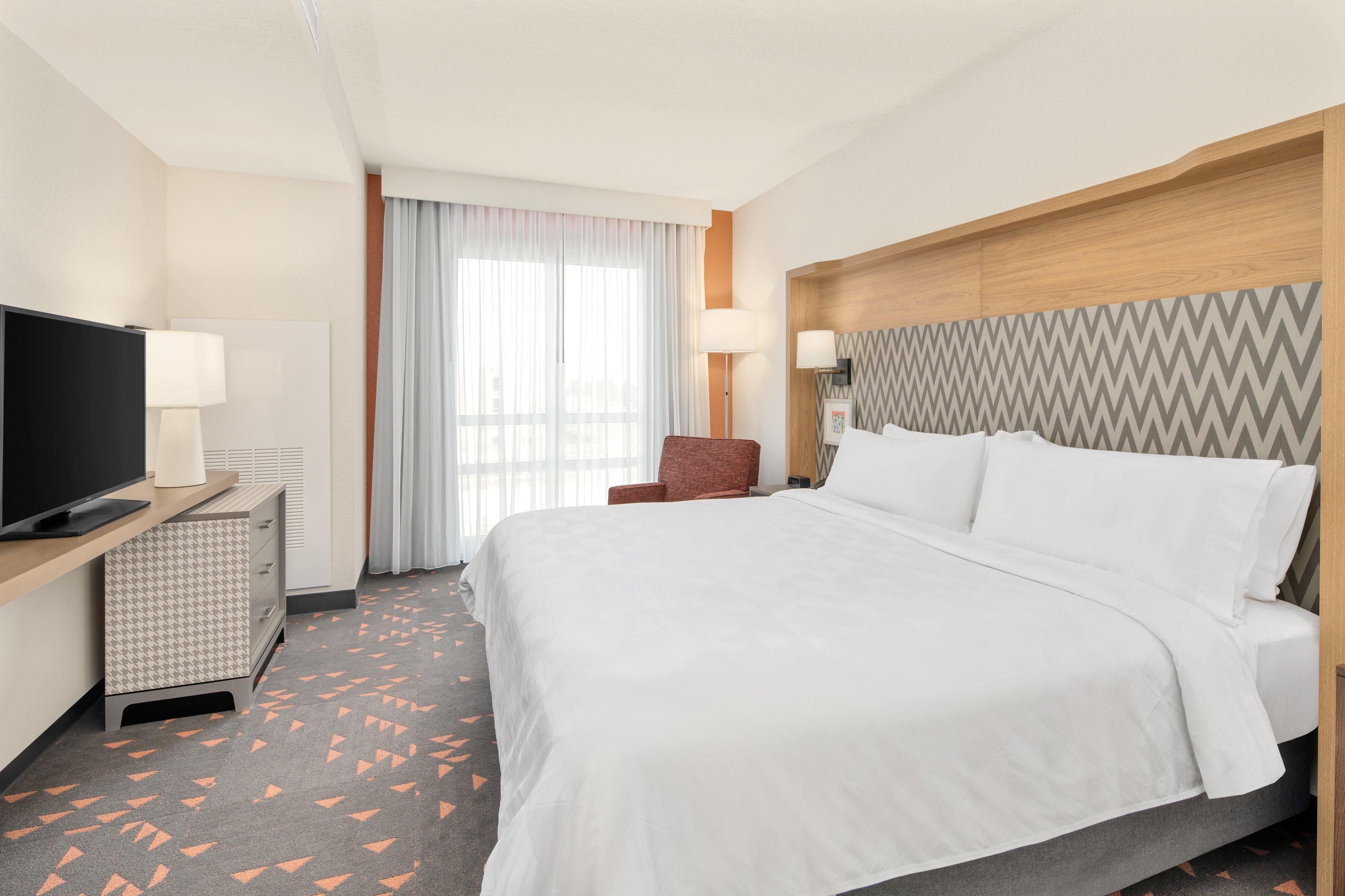 A hotel room with a bed, a TV, a desk with a lamp, a chair, and a patterned carpet.