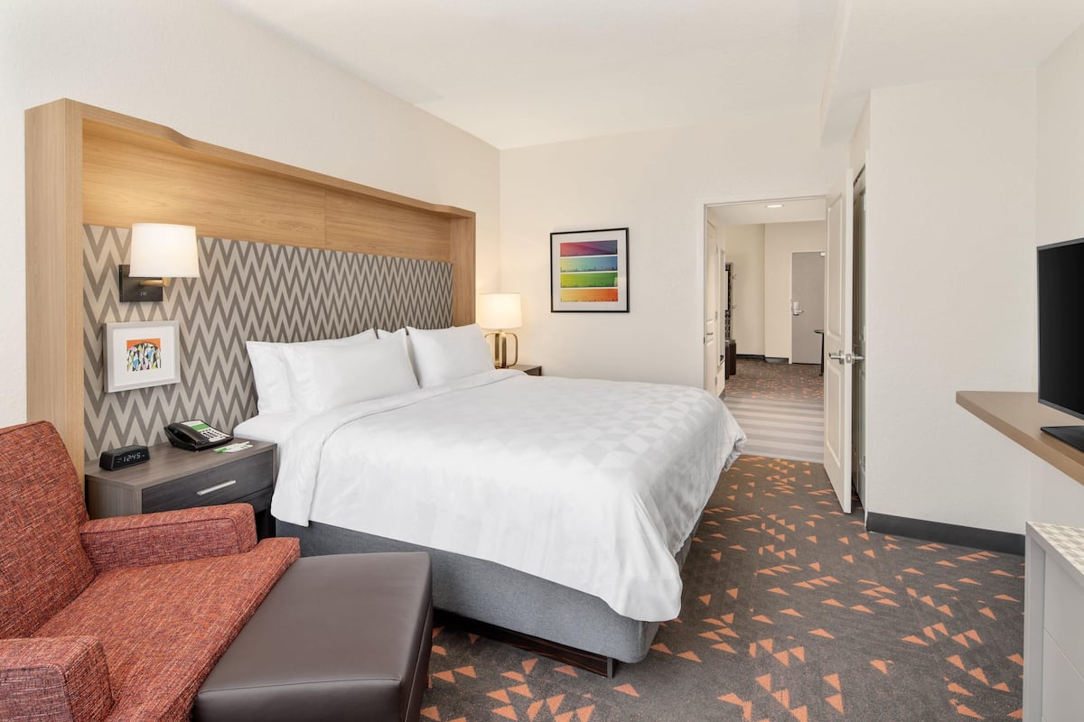 Ein Hotelzimmer mit Bett, Schreibtisch, Stuhl, Fernseher, Lampe, einem Bild an der Wand und einem Flur zu einem weiteren Zimmer.