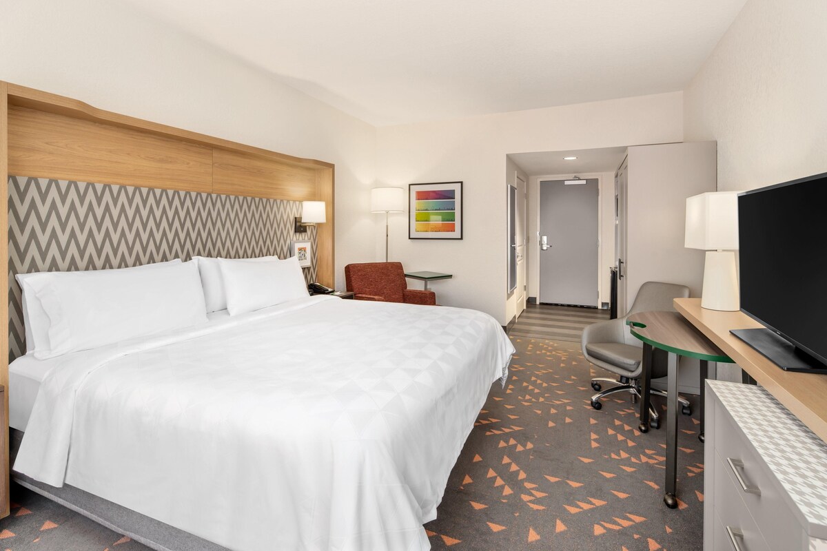 A hotel room with a large bed, a desk with a television, a chair, a desk lamp, and a wall-mounted lamp.