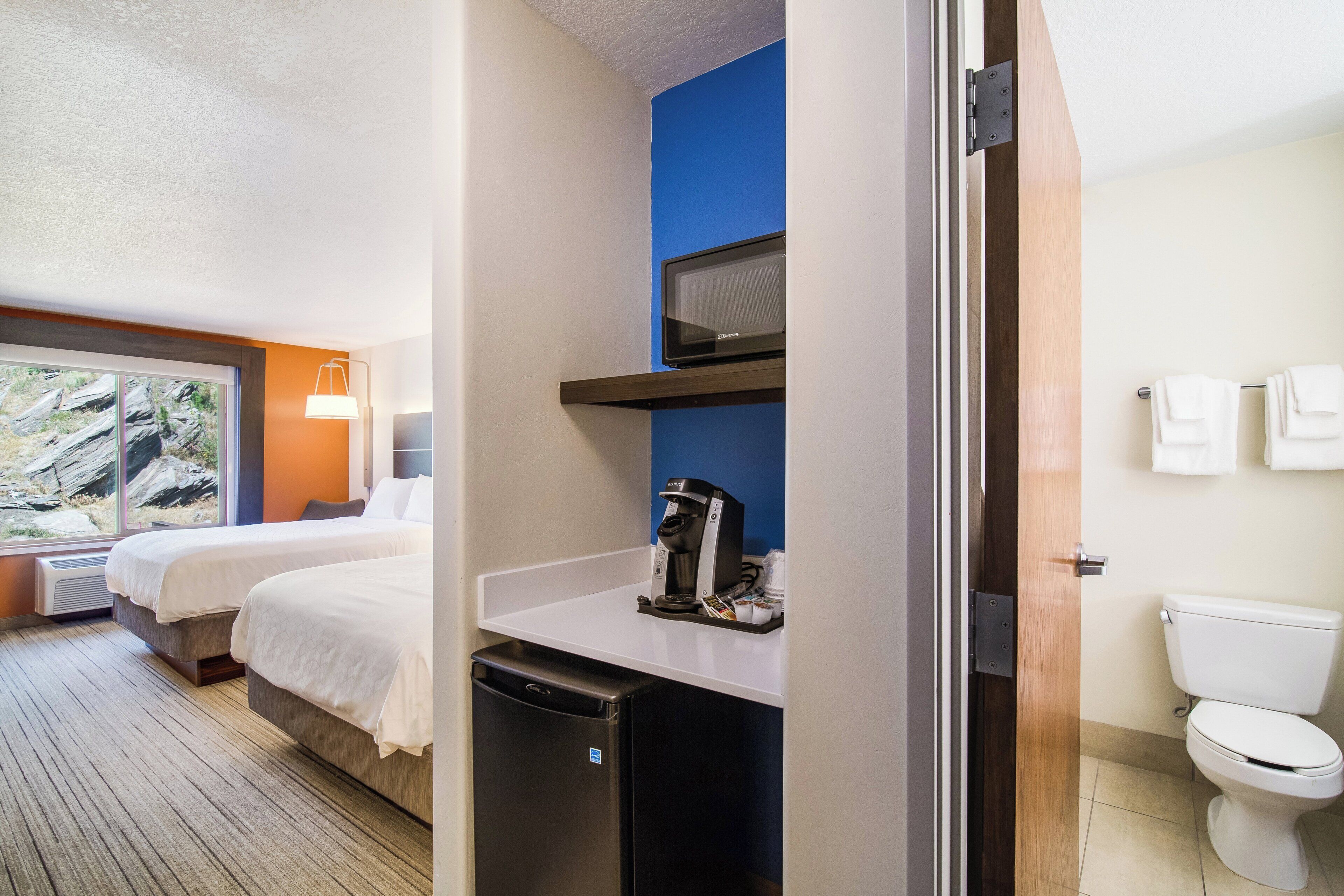 A hotel room with two beds, a small kitchenette, a microwave, a coffee maker, and a toilet.