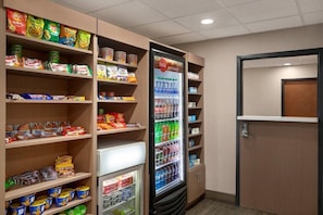 Ein Getränkeautomat, gefüllt mit verschiedenen Getränken und Snacks, umgeben von Regalen mit verpackten Waren.