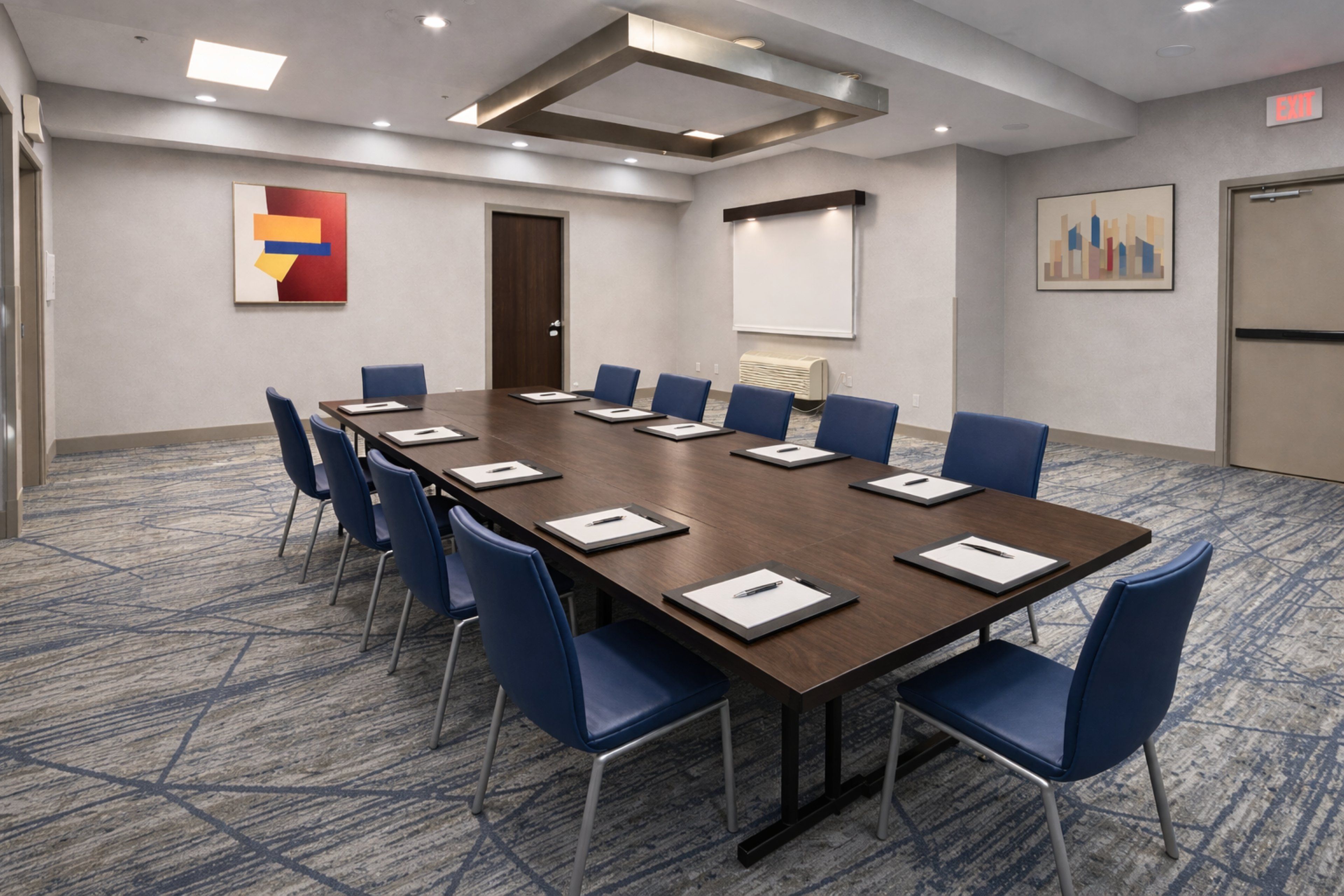 A conference room with a long table, blue chairs, and a projector screen.