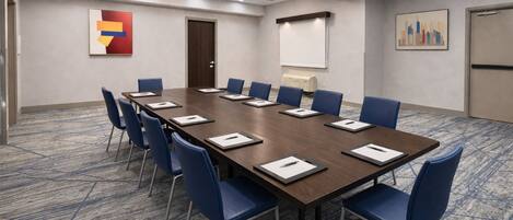 A conference room with a long table, blue chairs, and a projector screen.