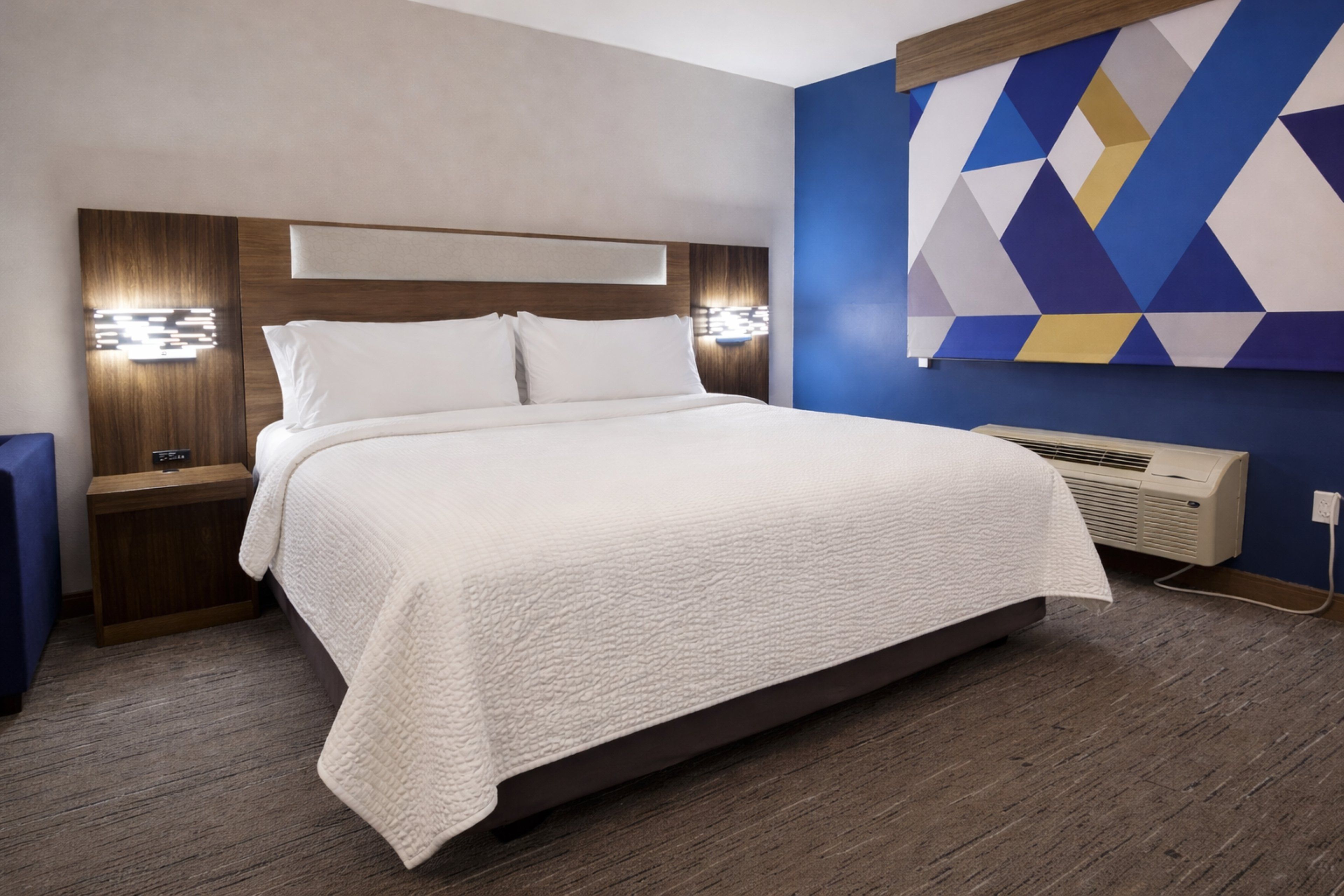 A hotel room with a large bed, wooden headboard, bedside lamps, and a wall-mounted air conditioner.
