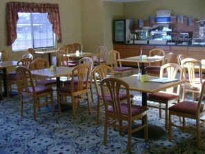 Zona pranzo con tavoli e sedie in legno, una finestra con tende e un angolo cottura con elettrodomestici.