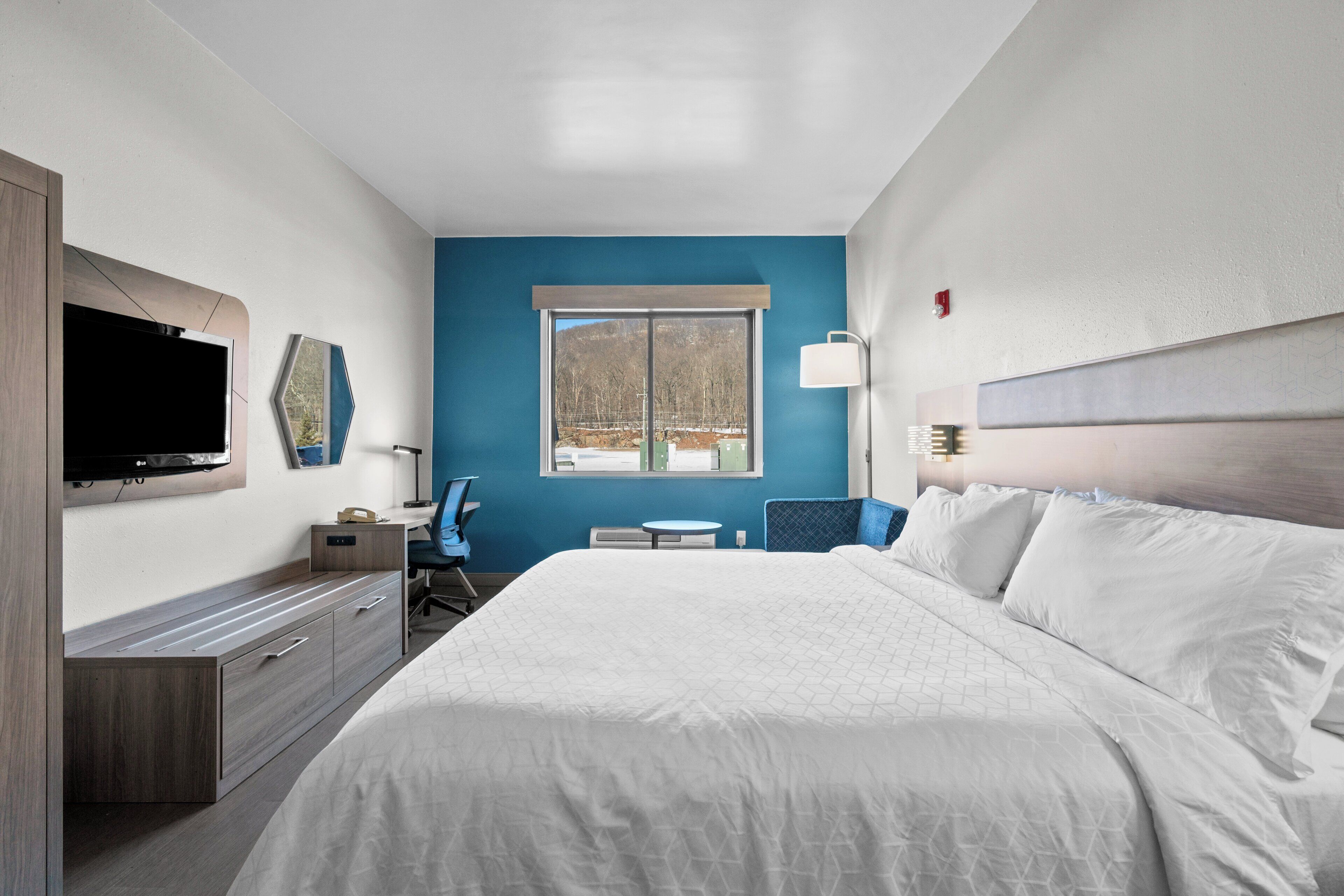 Quarto de hotel com uma cama grande, uma TV fixada na parede, uma mesa com cadeira e uma janela com vista para árvores.