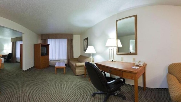 A hotel room with a desk, a chair, a sofa, a TV, a lamp, and a mirror.