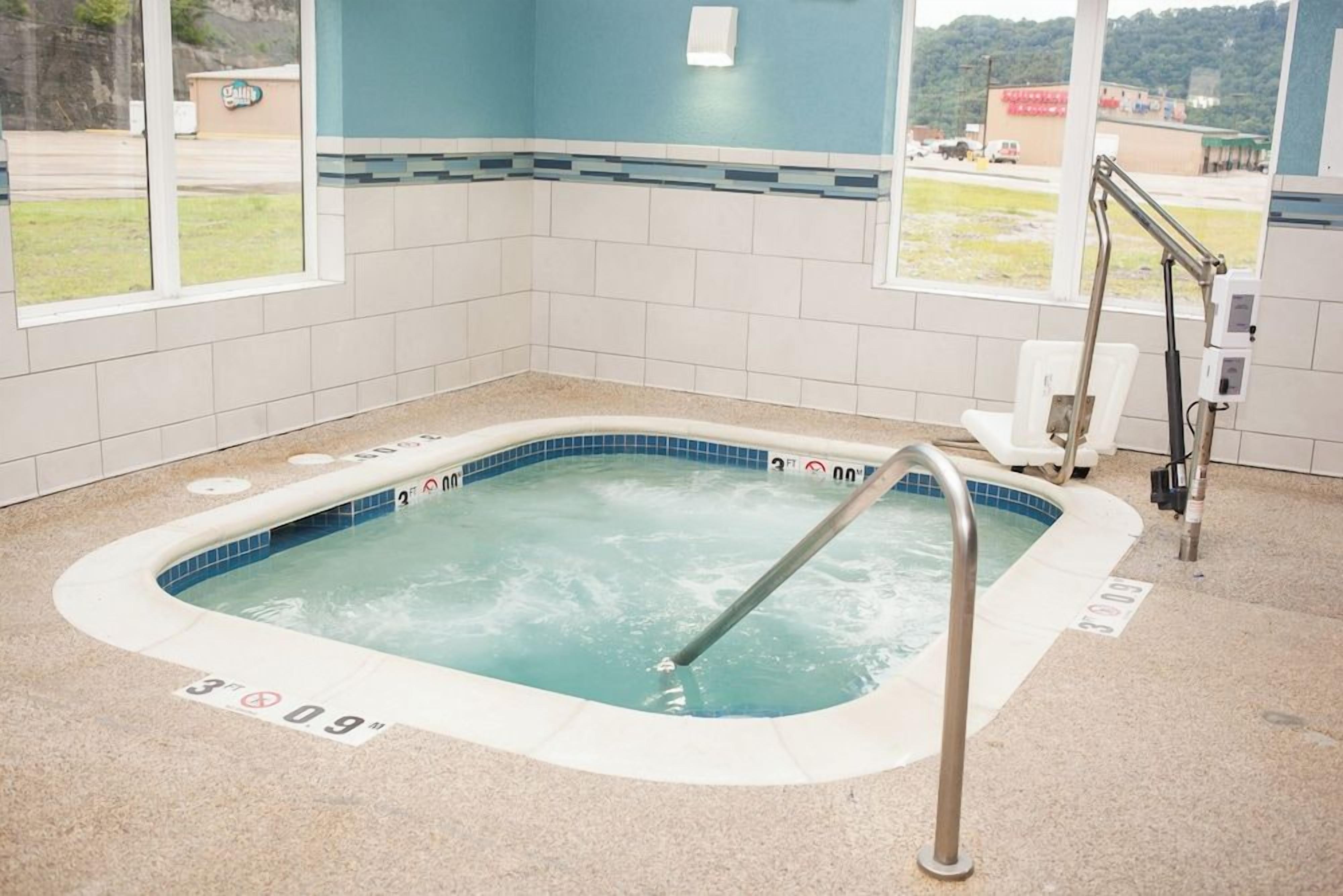 Un jacuzzi con una escalera metálica, suelo de baldosas y una ventana con vista a un estacionamiento y edificios.