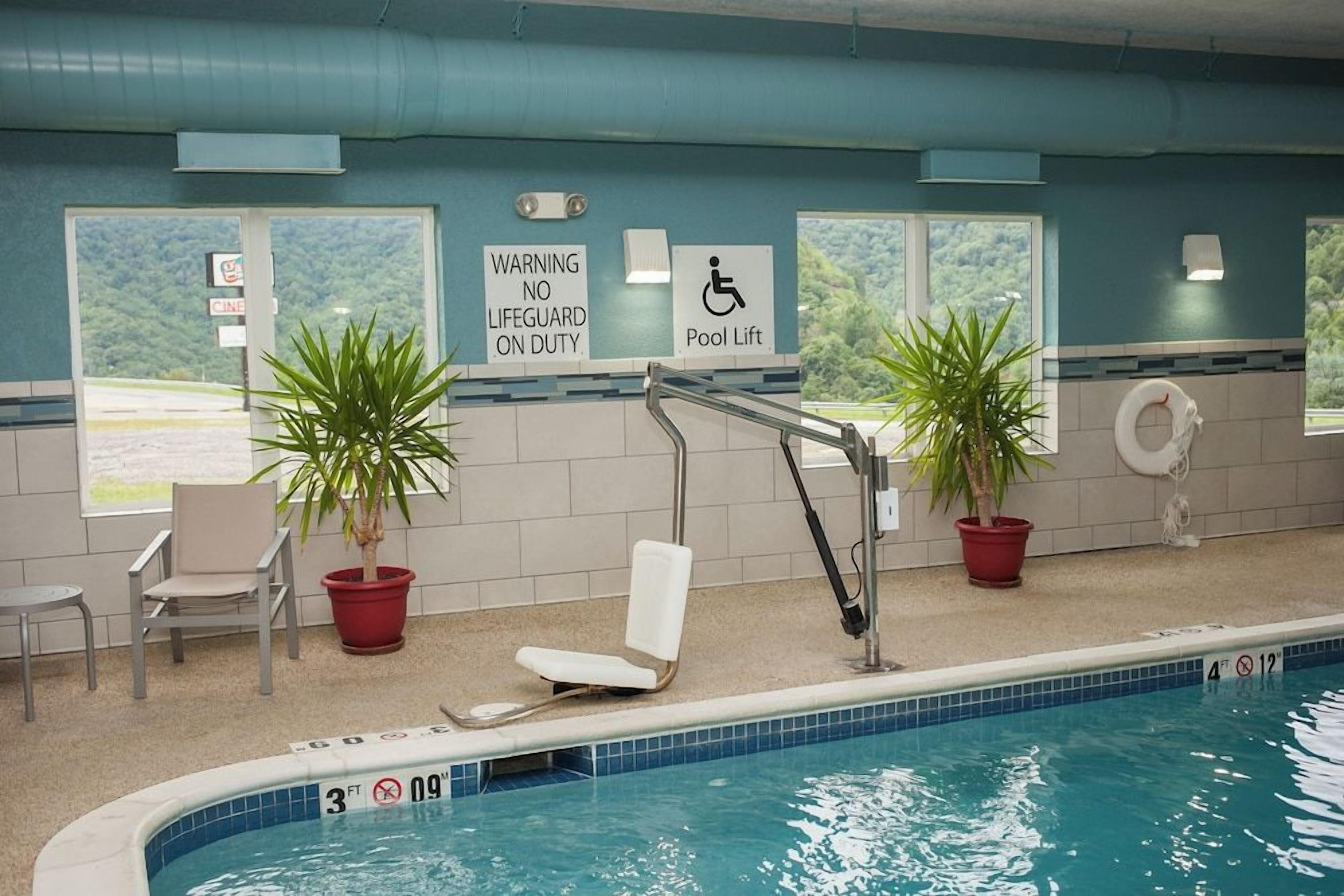 An indoor swimming pool with a lifeguard warning sign, a pool lift, and chairs.