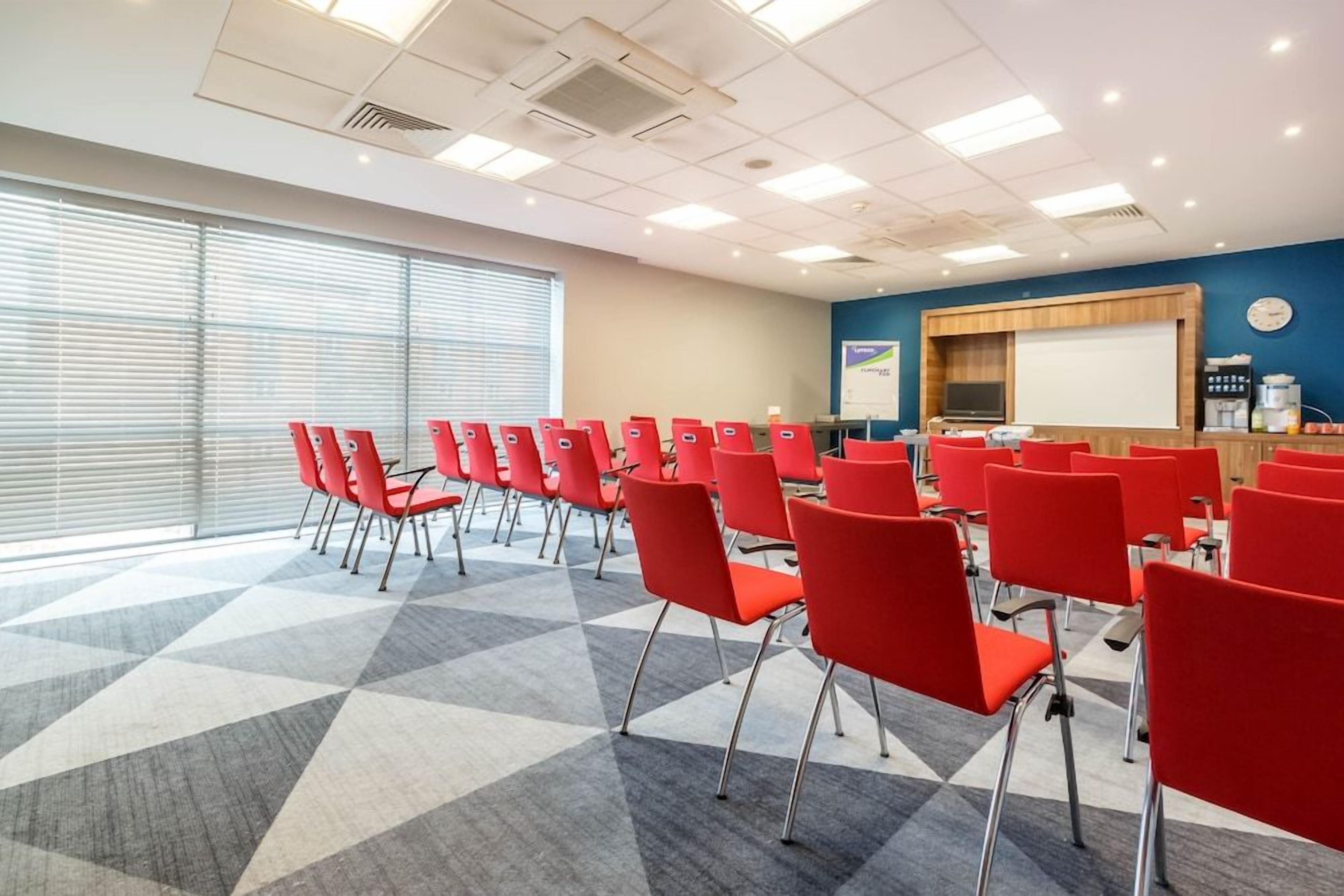A conference room with red chairs arranged in rows, a projector screen, and a presentation area with a coffee machine.