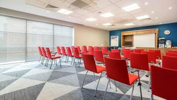 A conference room with red chairs arranged in rows, a projector screen, and a presentation area with a coffee machine.
