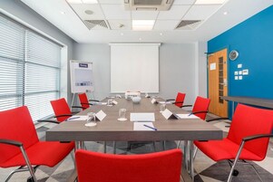 Un salón de conferencias con una gran mesa, sillas rojas, una pizarra y una pantalla de proyector.