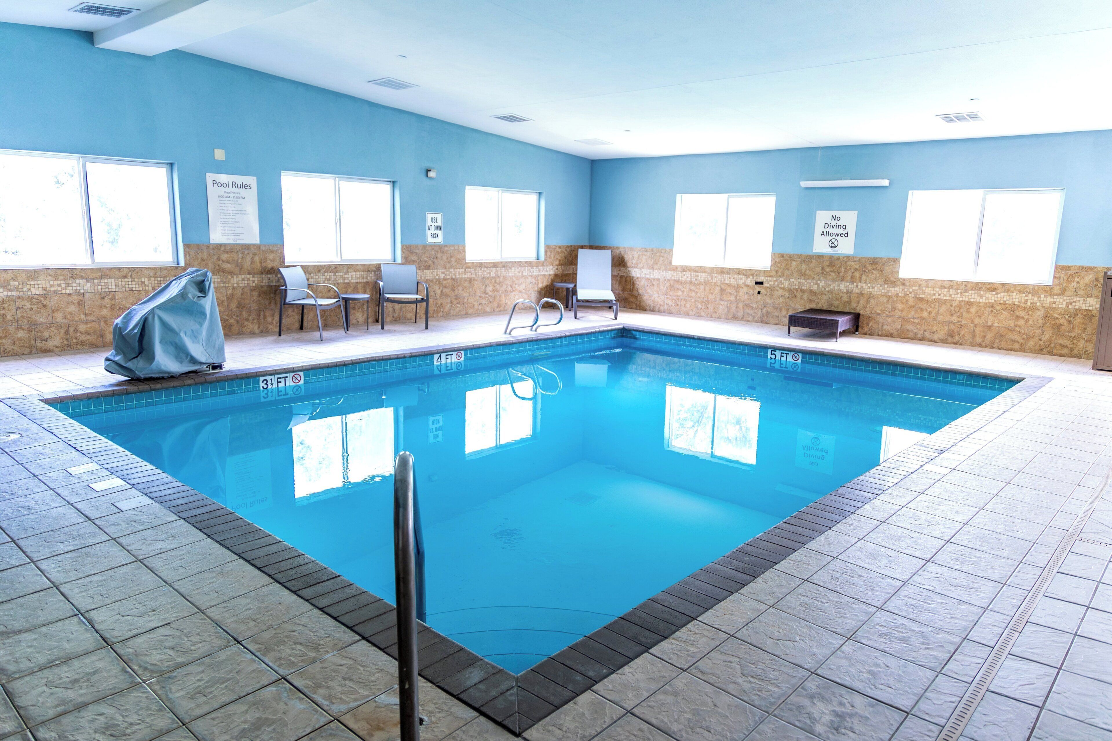 An indoor swimming pool with clear pool rules and no diving allowed signs, surrounded by tiled walls and a tiled floor.