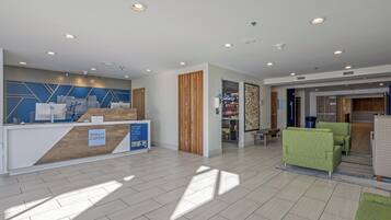 A hotel reception area with a modern design, including a reception desk, a seating area with green sofas, and a visible storage room in the background.