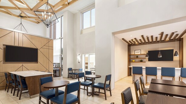 A modern dining area with wooden tables and blue chairs, a large flat-screen TV, and a kitchen area with a wooden ceiling.