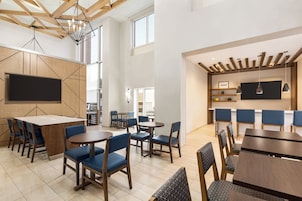 A modern dining area with wooden tables and blue chairs, a large flat-screen TV, and a kitchen area with a wooden ceiling.