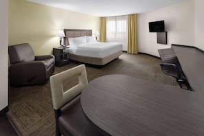 A hotel room with a bed, two chairs, a desk, a TV, and a window with curtains.