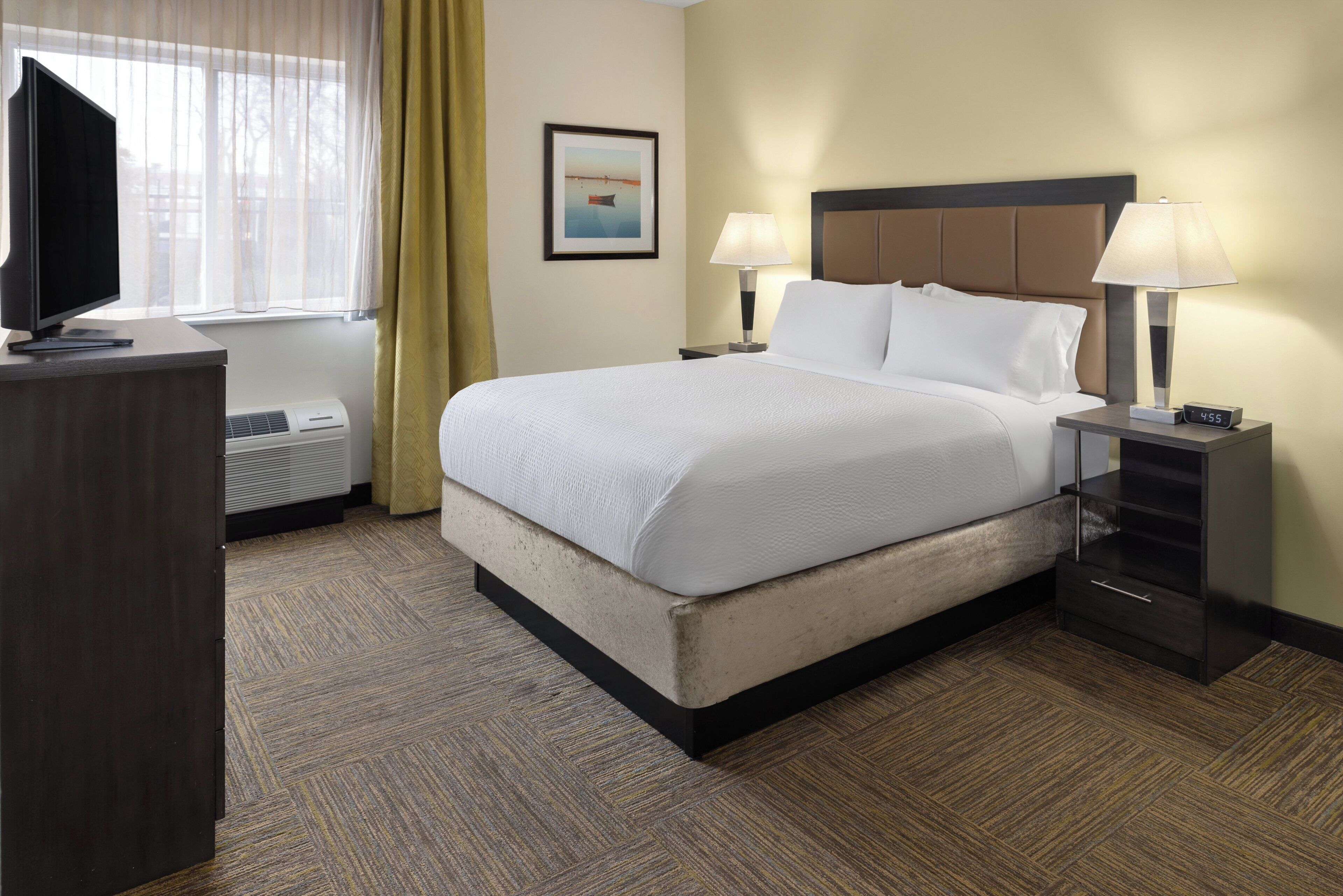 A hotel room with a bed, bedside lamps, a TV, a framed picture on the wall, and a window with curtains.