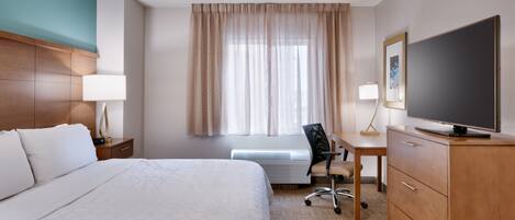 Habitación de hotel con cama, escritorio, silla, televisor y ventana con cortinas.